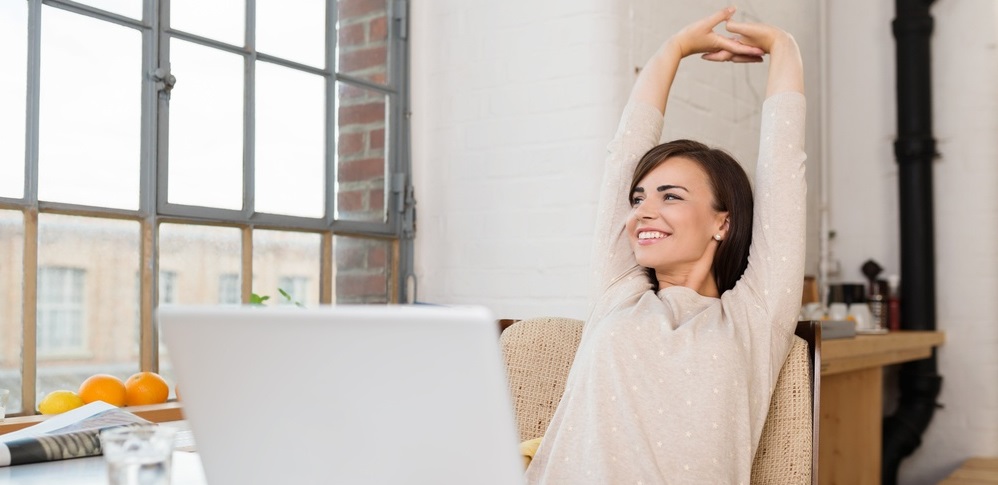 Happy relaxed young woman sitting in her kitchen with a laptop in front of her stretching her arms above her head and looking out of the window with a smile